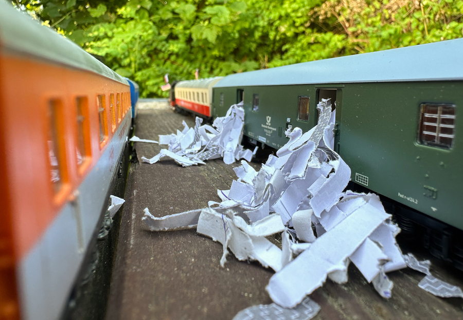 Two model trains are parked on the platform, shredded paper falling out of the open doors from the train on the opposite side.
