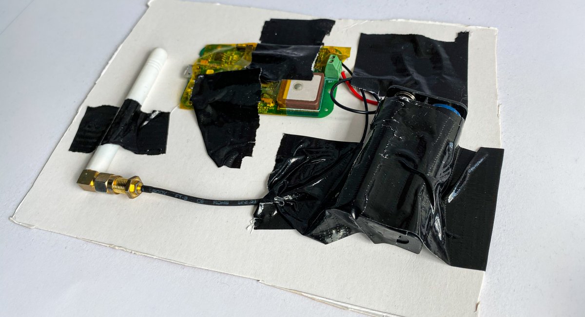 GPS-Tracker, Batterie-Halterung und Antenne mit abgerupftem Gaffertape auf eine schief ausgeschnittene Pappe geklebt